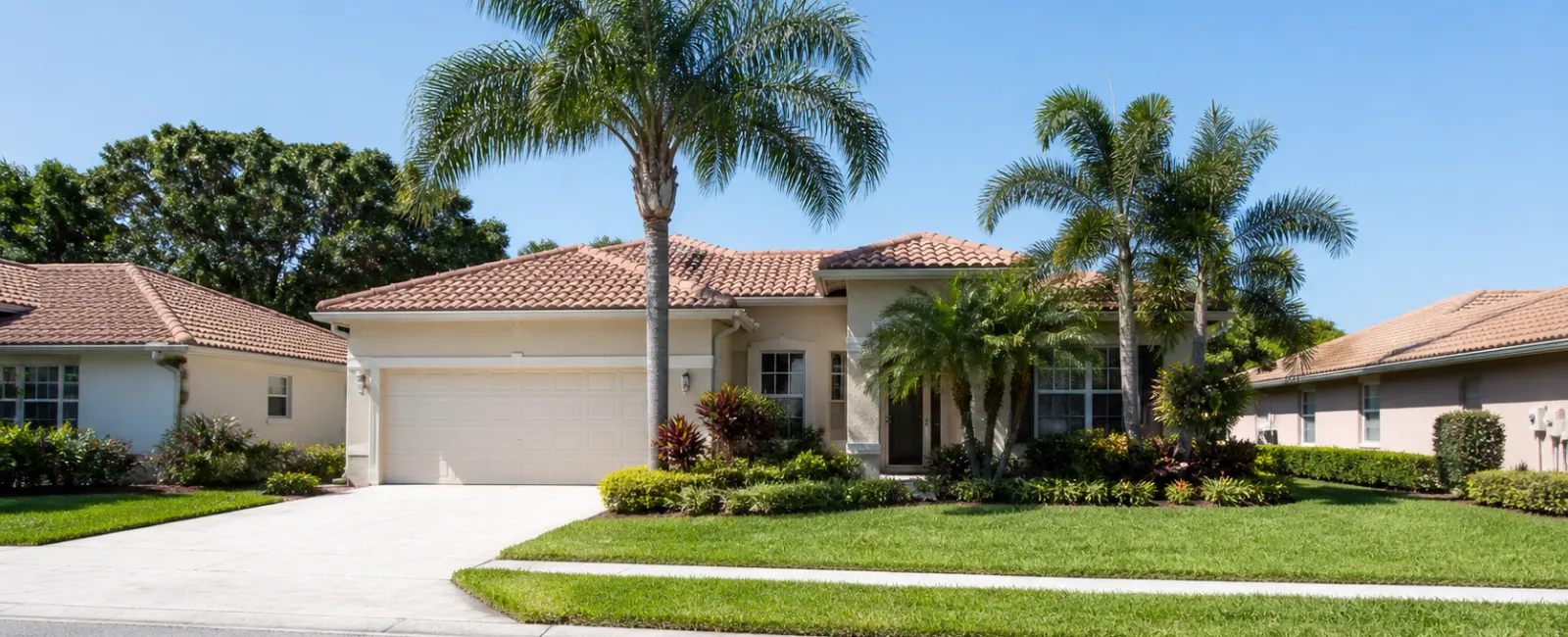 Single-story suburban house with tiled roof, palm trees, and manicured lawn under a clear blue sky.