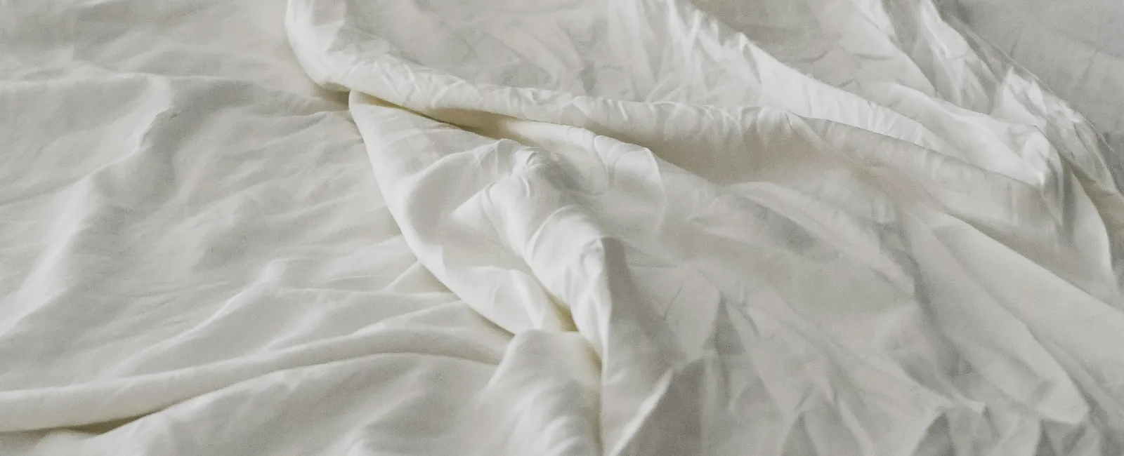 Close-up of a rumpled white bed sheet and pillow on a neatly made bed with soft natural lighting.