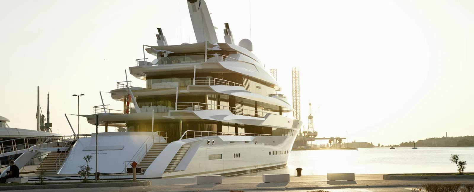 Luxury white yacht docked at harbor with calm water and golden sunset in the background.