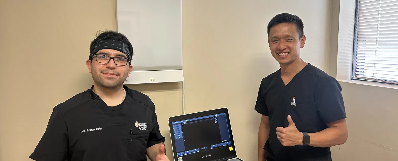 Two male medical professionals in black scrubs giving thumbs up next to an ultrasound machine in a clinic room.