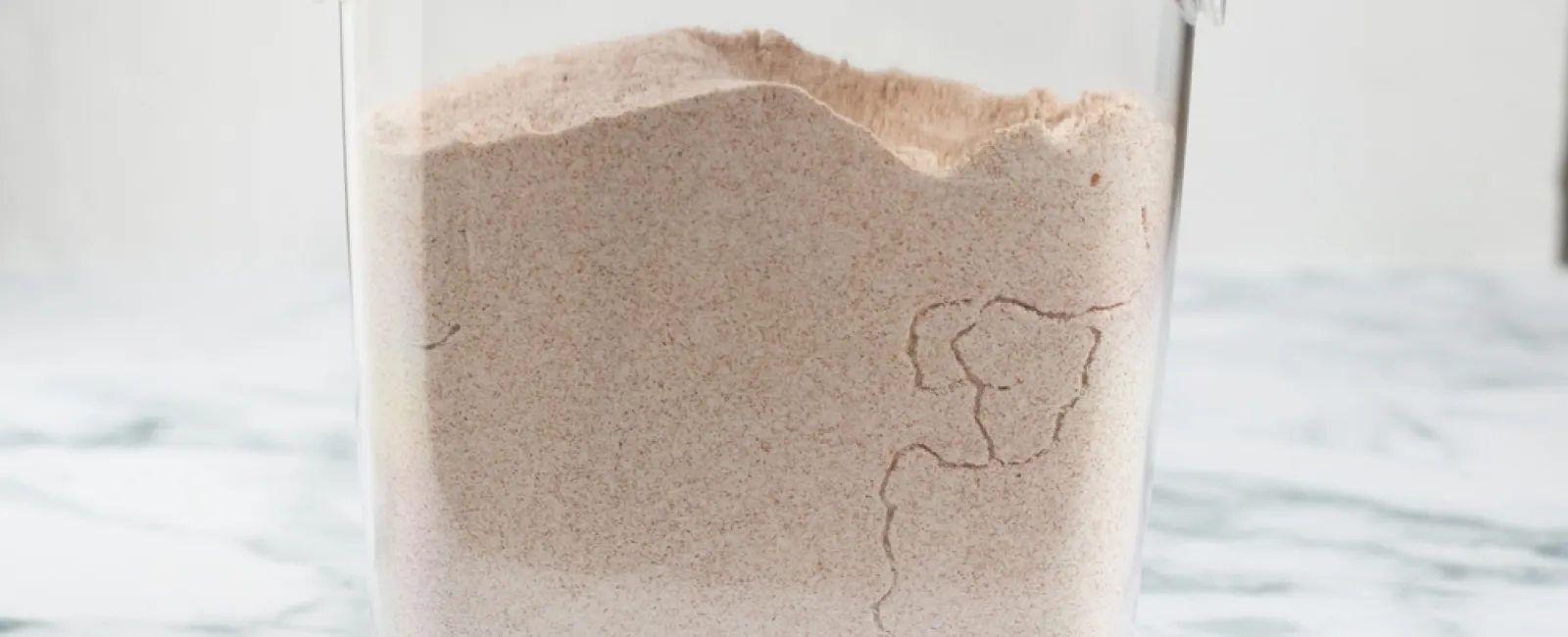 Clear plastic container filled with whole wheat flour on a marble countertop with a white lid.