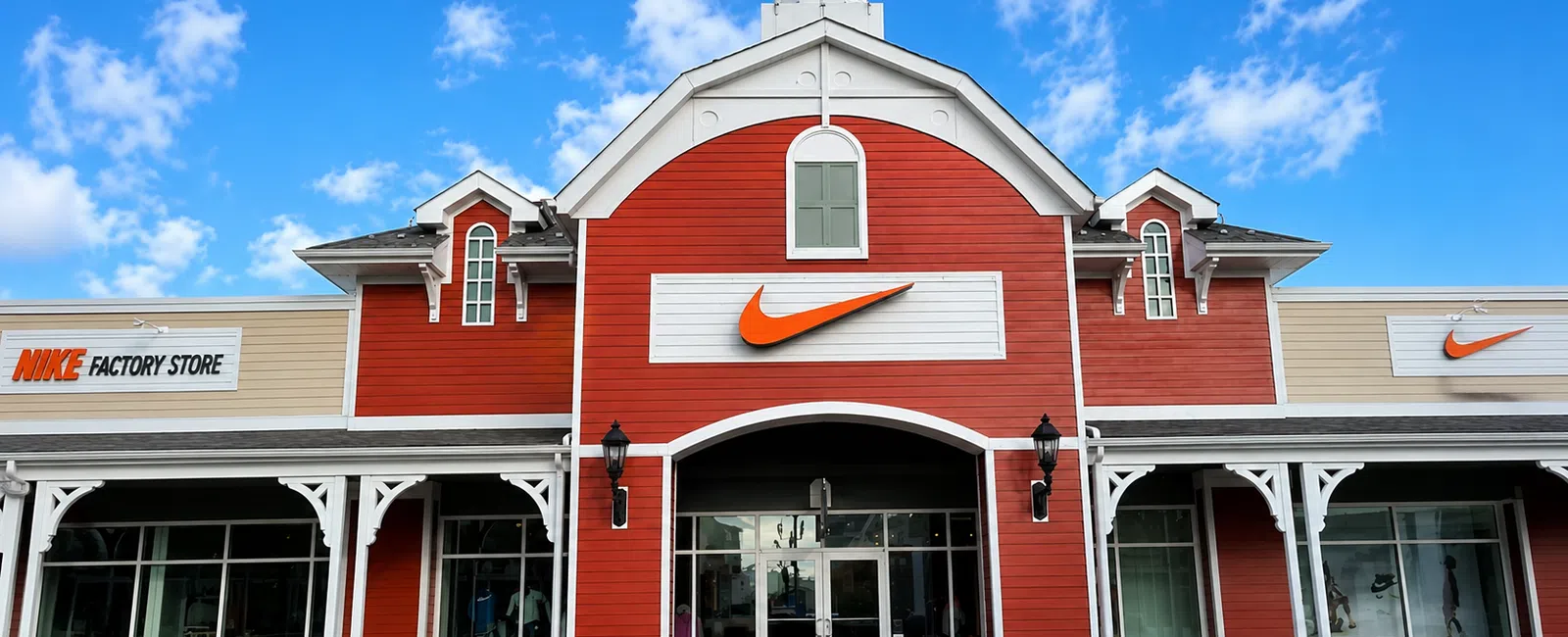 Red and beige Nike Factory Store building with large orange swoosh logos under a bright blue sky.
