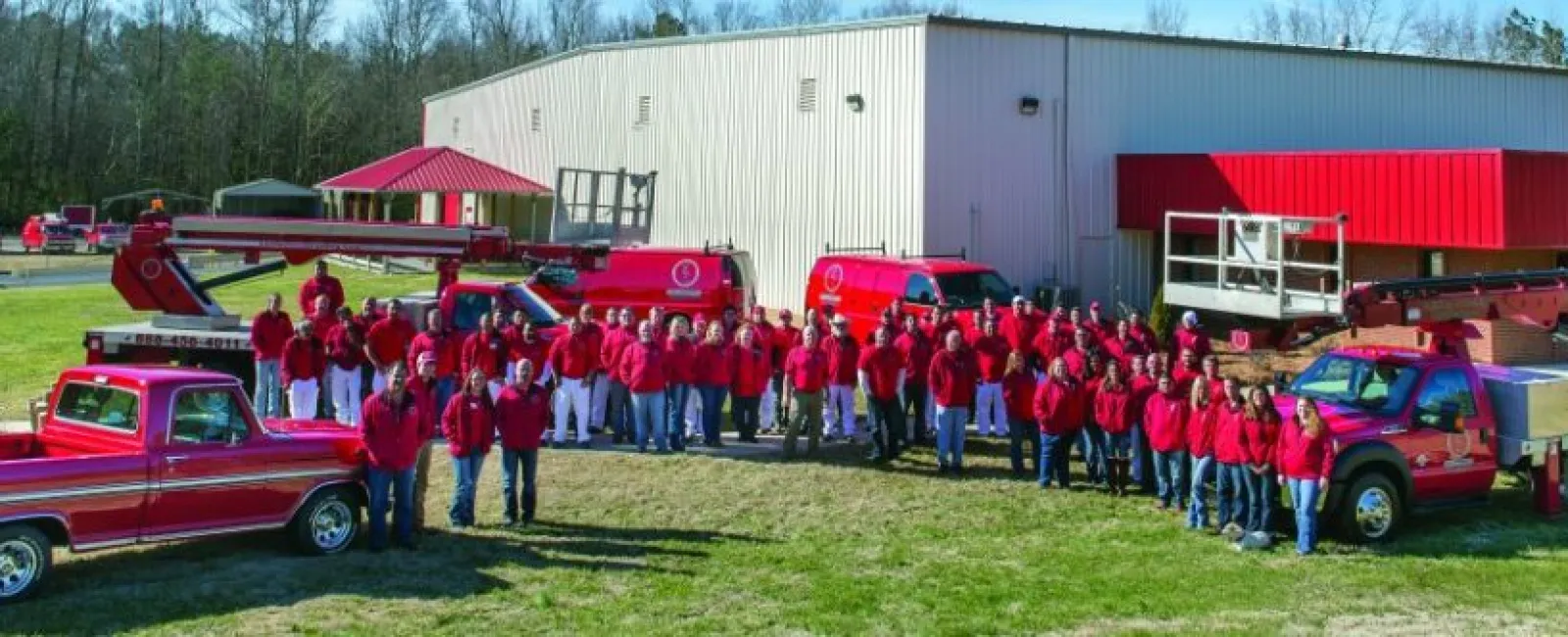Large group of people wearing red shirts posing in front of red vehicles and a warehouse under clear sky.