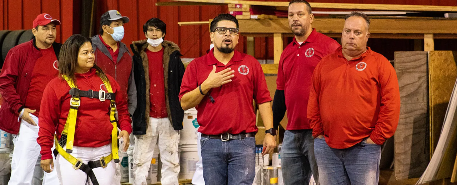 Group of workers in red shirts in a warehouse, one wearing a safety harness and others listening attentively.