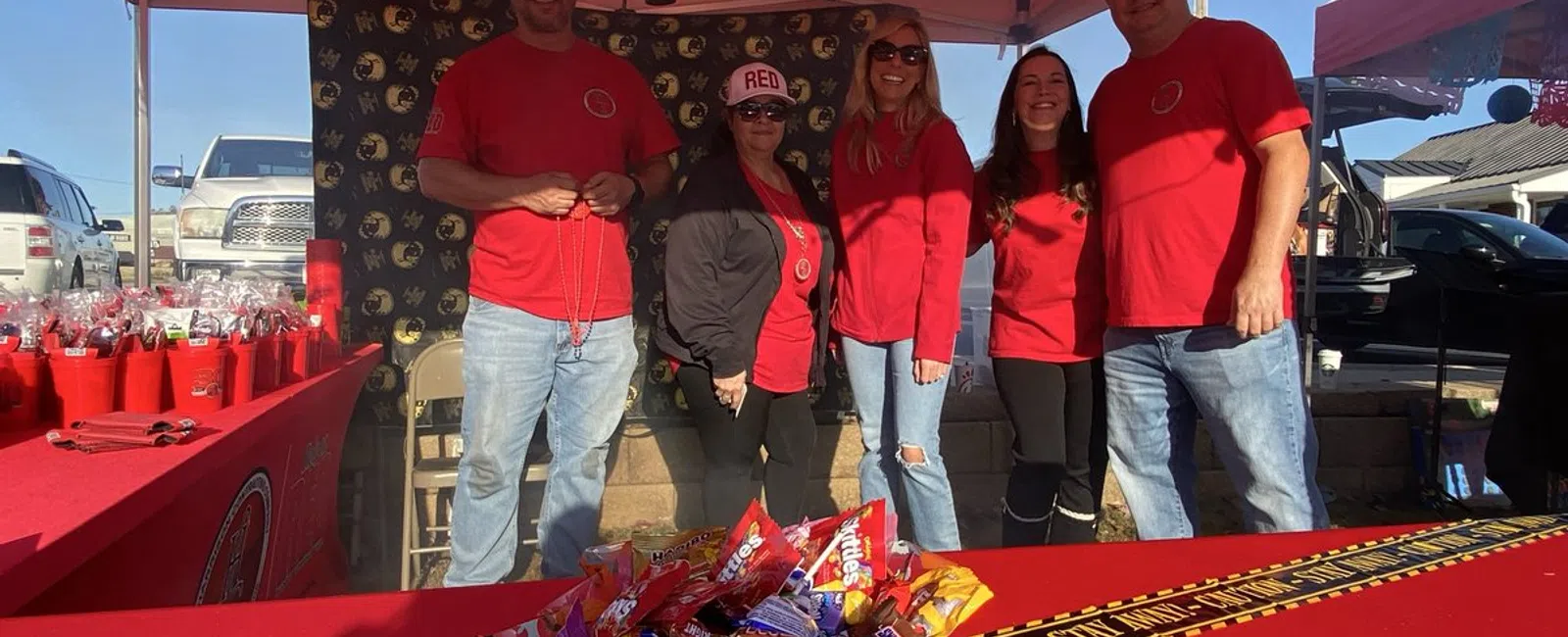 Group of five people in red shirts under a tent with Halloween decorations and candy on a table with caution tape.