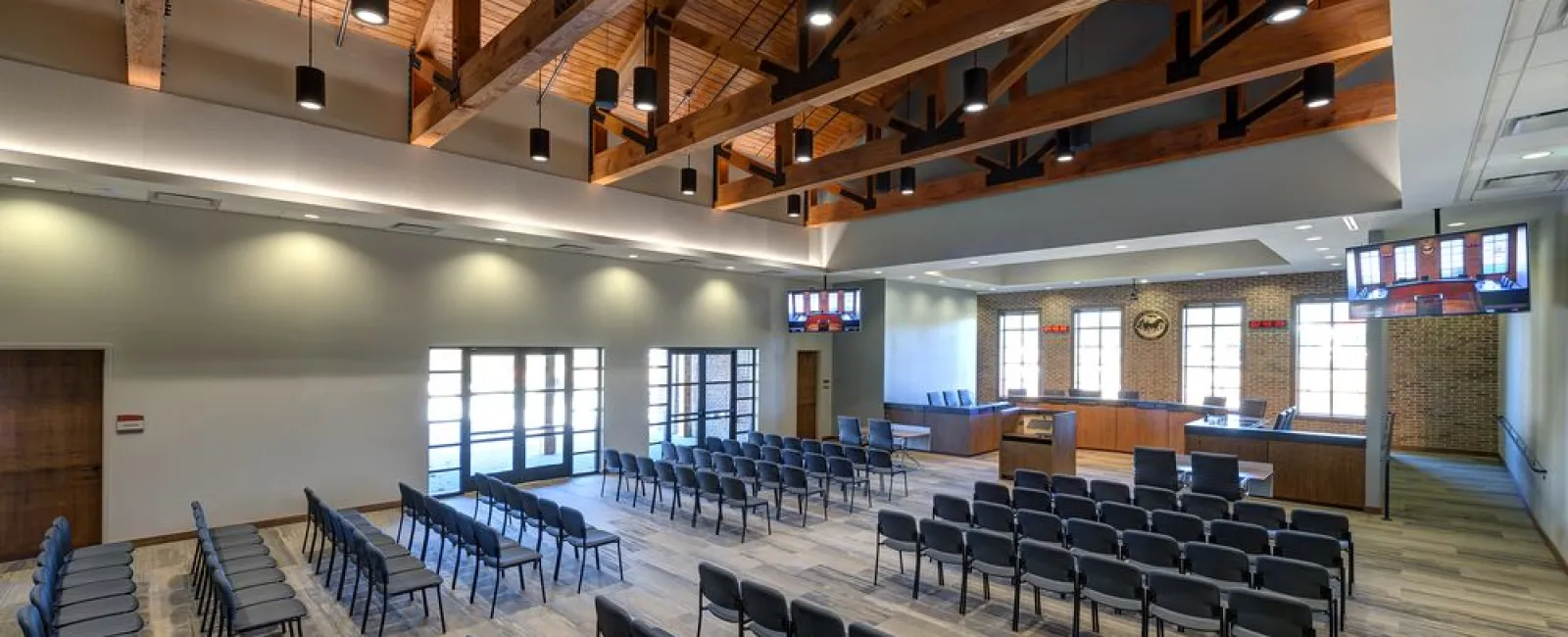 Modern courtroom with wooden ceiling beams, rows of black chairs, and a judge's bench with monitors