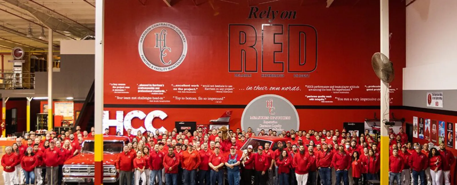 Large group of employees wearing red in front of a red wall with motivational text and company logos in a warehouse setting