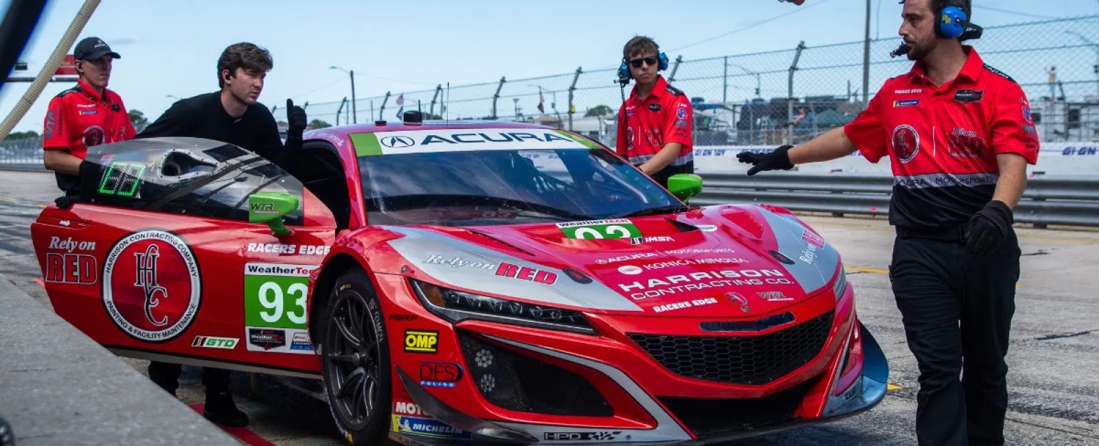 Red Acura race car number 93 in pit stop with crew members in red uniforms on racetrack.