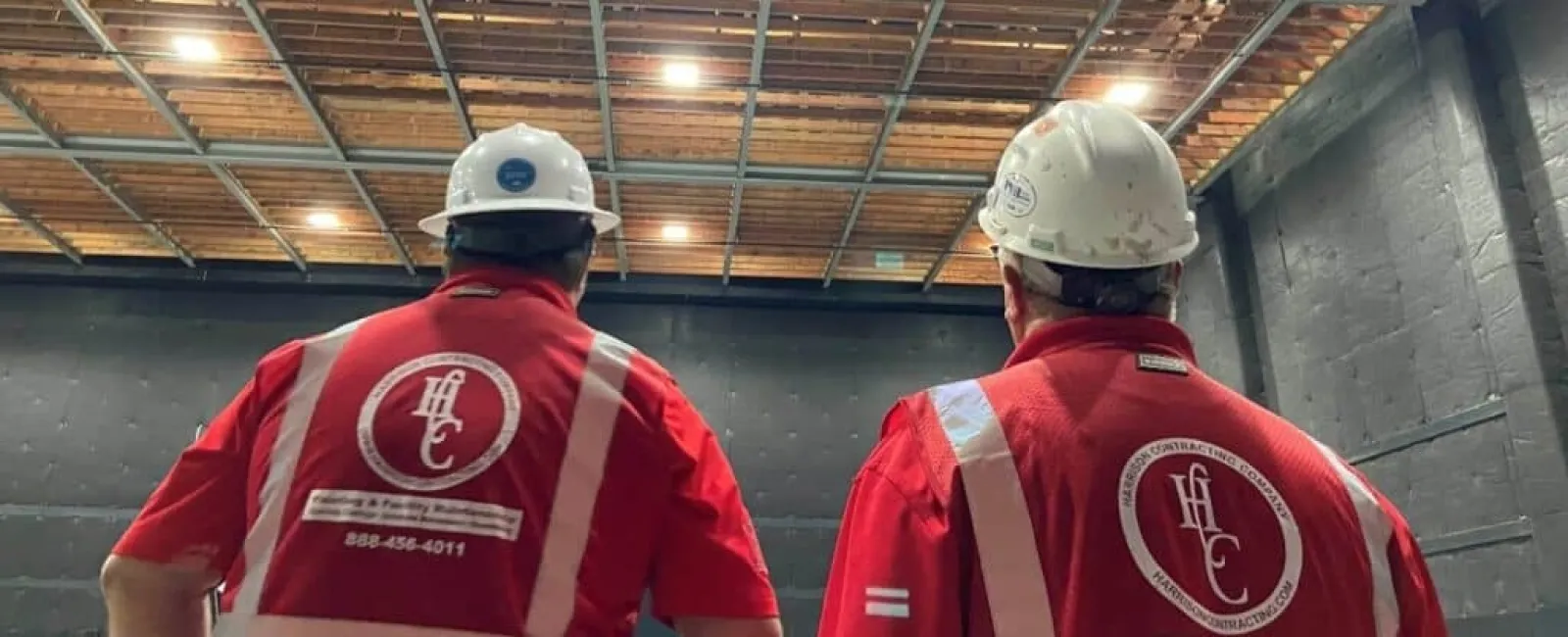 Two workers in white helmets and red safety vests inside a large industrial space with exposed ceiling beams.
