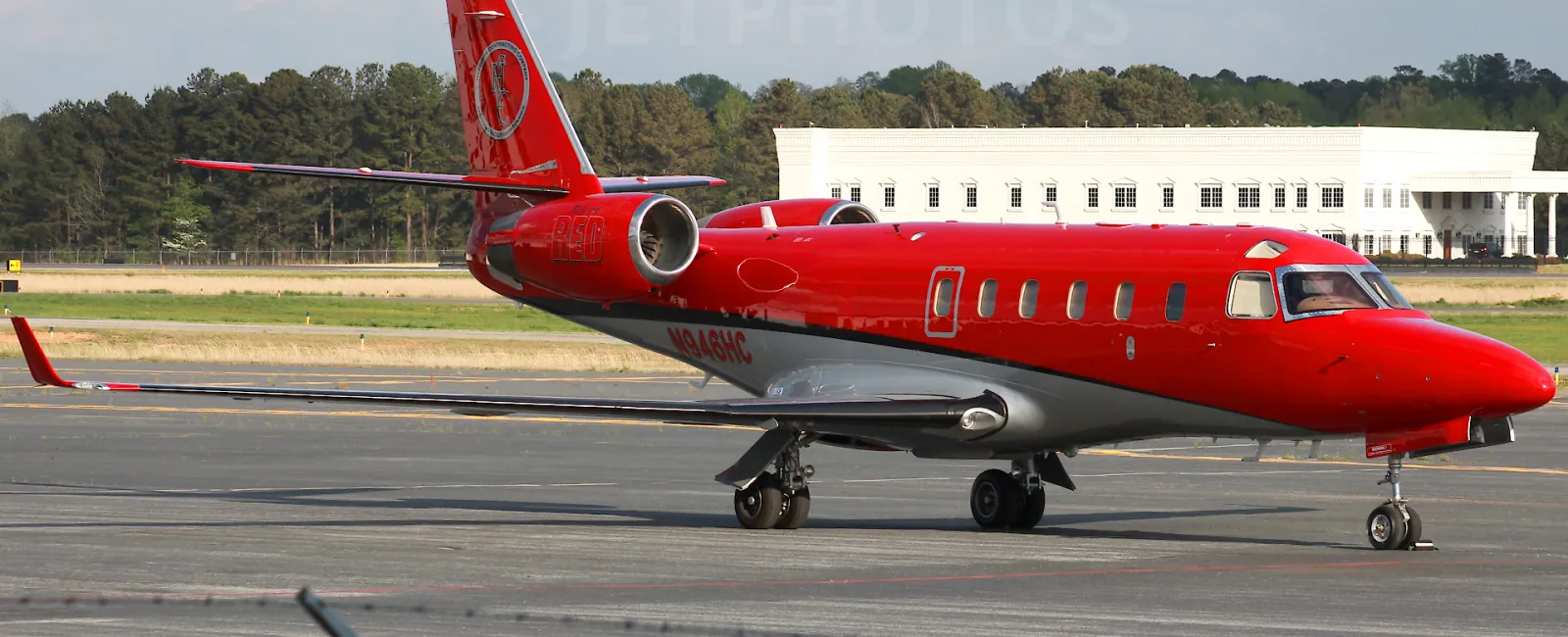 Red and white private jet parked on runway with building and trees in the background on a cloudy day