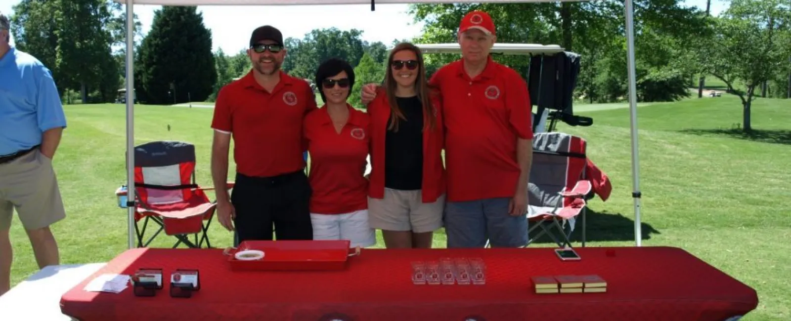 Four people under company tent with a branded tablecloth at an outdoor event on a sunny day.