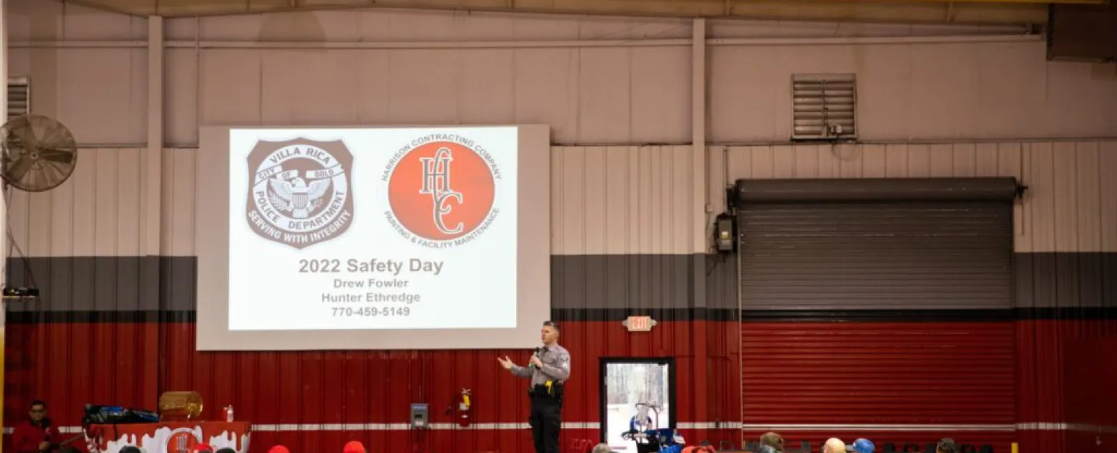Speaker presenting 2022 Safety Day to a large indoor audience wearing red and yellow safety gear in a warehouse.