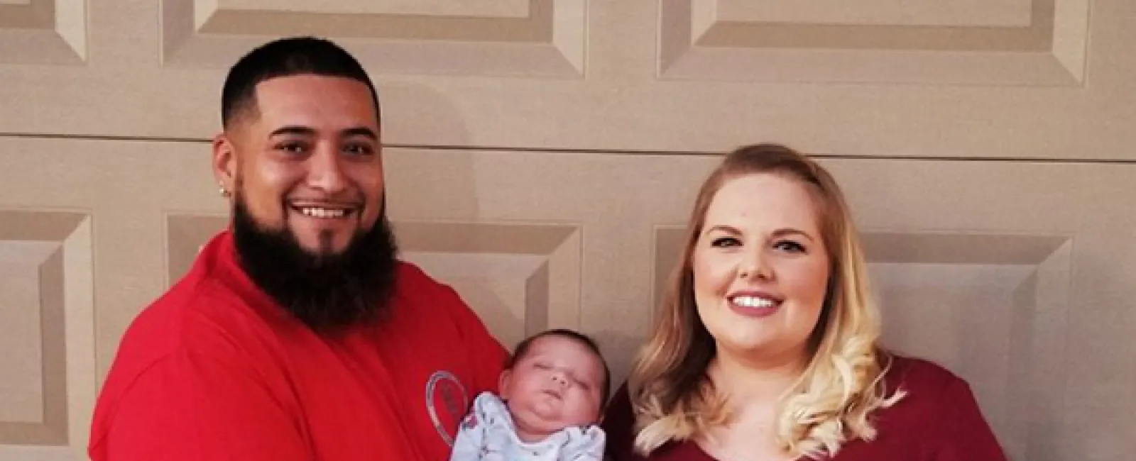 Smiling couple standing in front of a garage door holding a newborn baby, all dressed in red and white clothes.