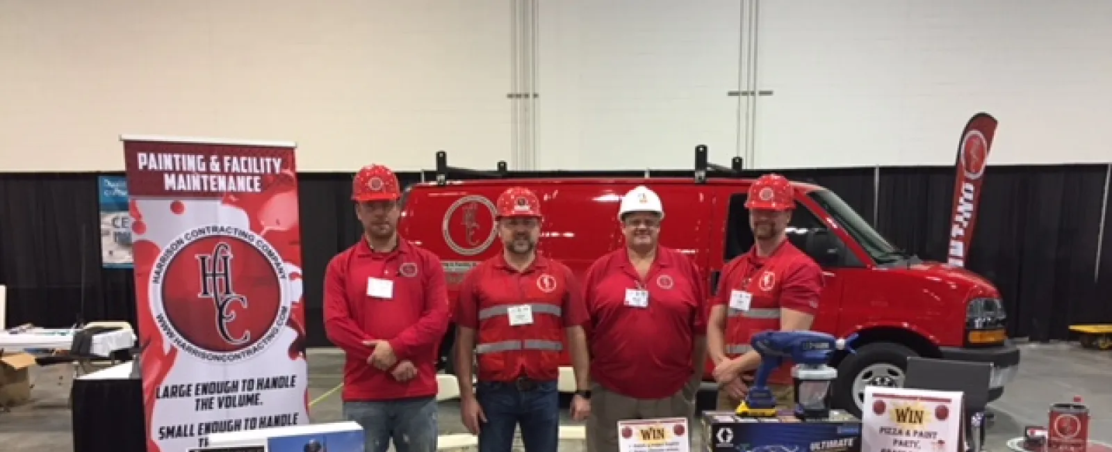 Four men wearing red shirts and hard hats stand behind a red table with promotional items and a van at a trade show booth.