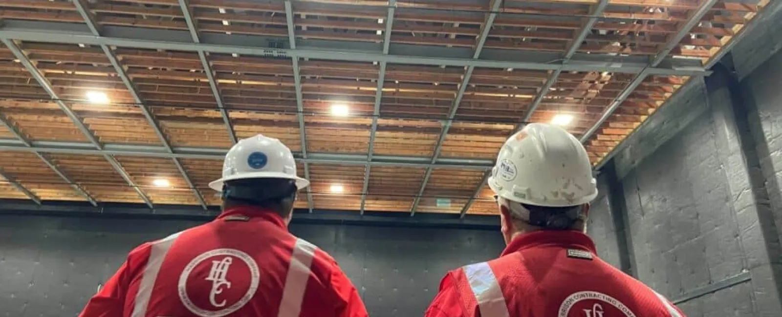 Two construction workers in red safety vests and white helmets inspecting an unfinished indoor ceiling.