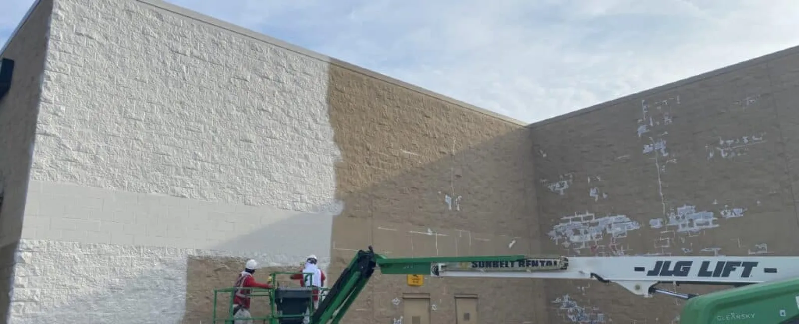 Workers on a green JLG lift painting a large exterior beige wall white under a blue sky.