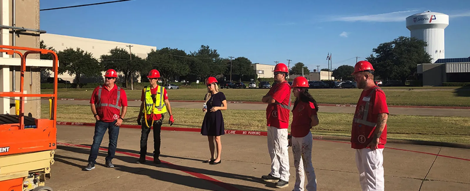 Group of workers and a supervisor wearing red helmets and safety gear standing outdoors near a scissor lift on a sunny day