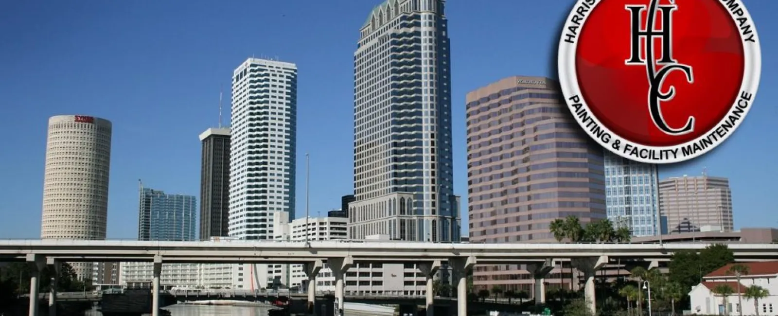City skyline with modern high-rise buildings and a river in the foreground under a clear blue sky.