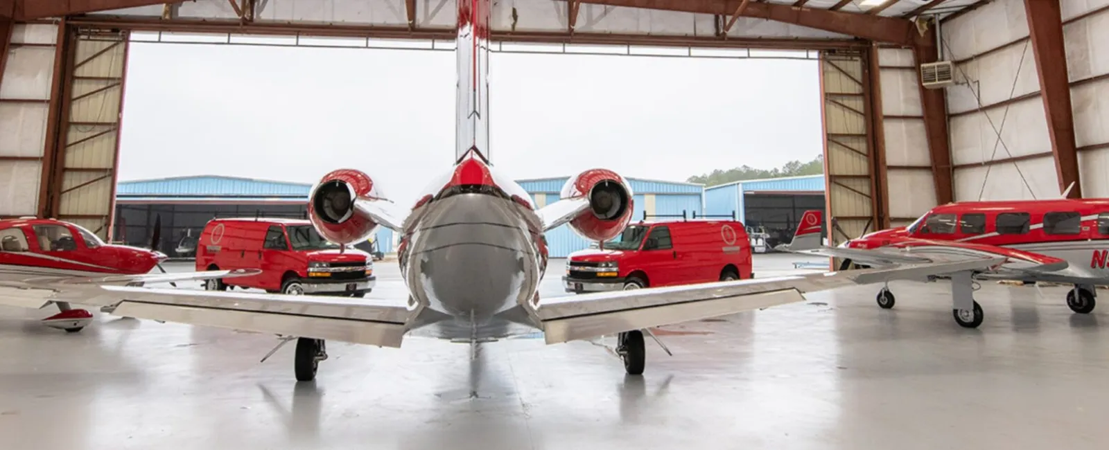 Front view of a private jet in a hangar flanked by two red single-engine airplanes and red service vans.