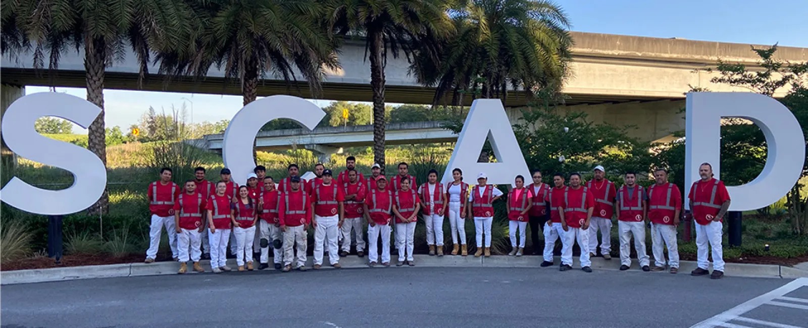 Group of people in red vests and white pants standing in front of large SCAD letters with palm trees and overpass behind.