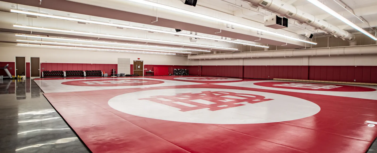 Indoor wrestling gym with red mats featuring large white circles and a logo, bright overhead lighting.