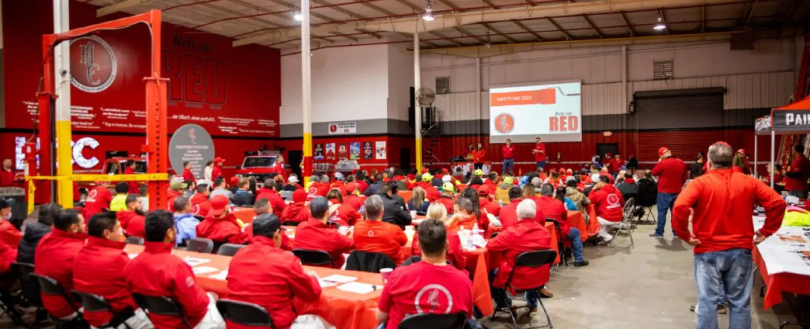 Large group of workers in red uniforms attending safety training session in a spacious industrial warehouse.
