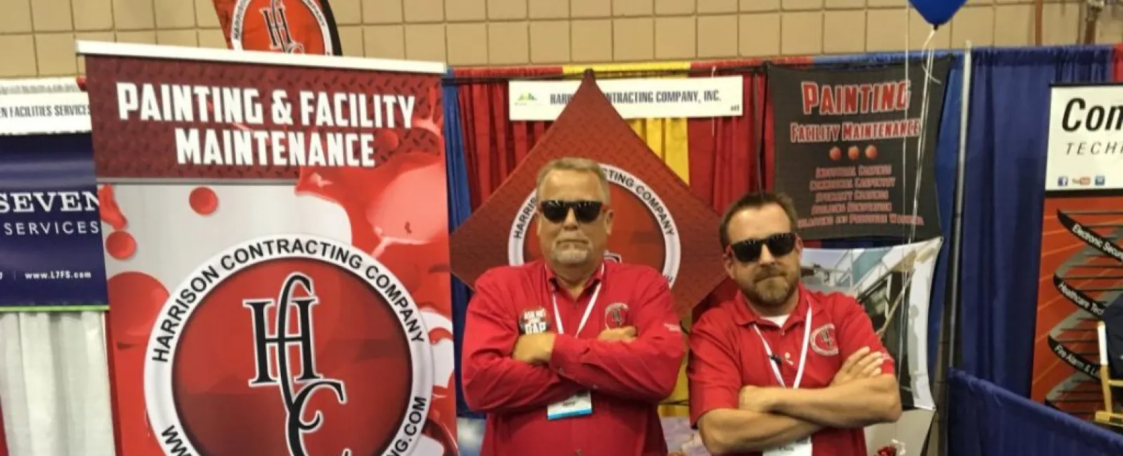 Two men in red shirts with sunglasses stand in front of Harrison Contracting Company booth at a trade show.