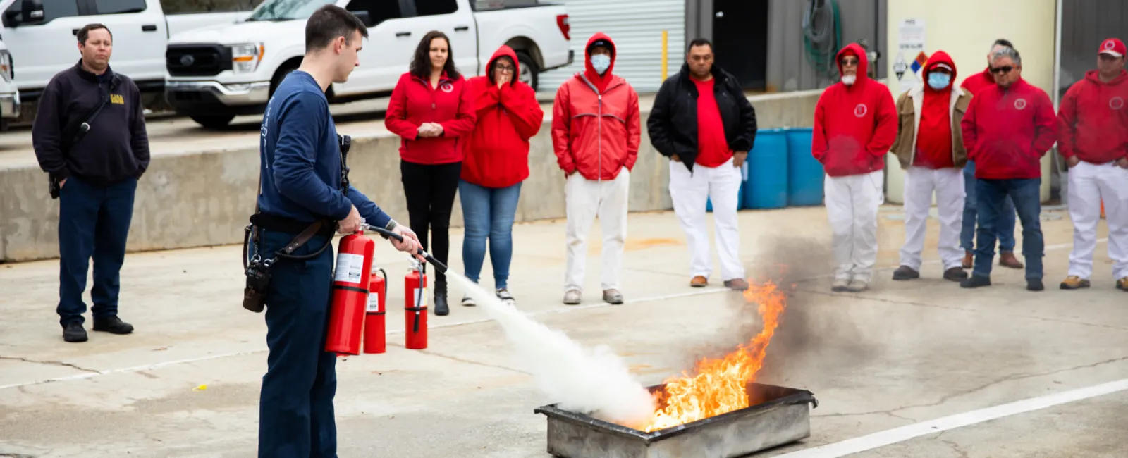 Person using fire extinguisher to put out a controlled fire during outdoor safety training with group observing.