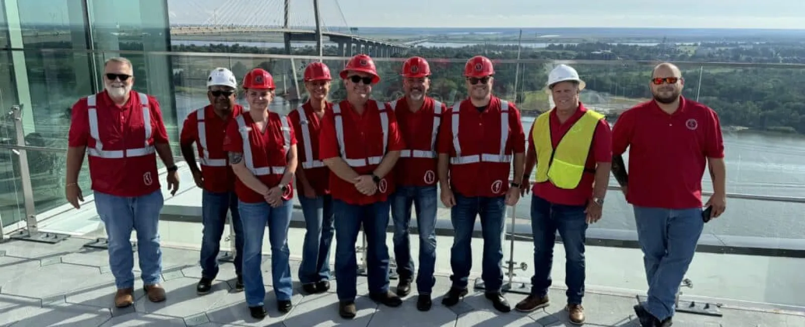 Group of construction workers wearing red safety vests and helmets standing on rooftop with bridge and river view