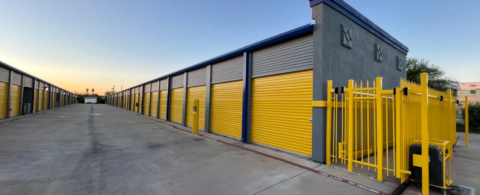 Wide view of a storage facility with yellow roll-up doors and gated entrance under a clear sky at sunset.