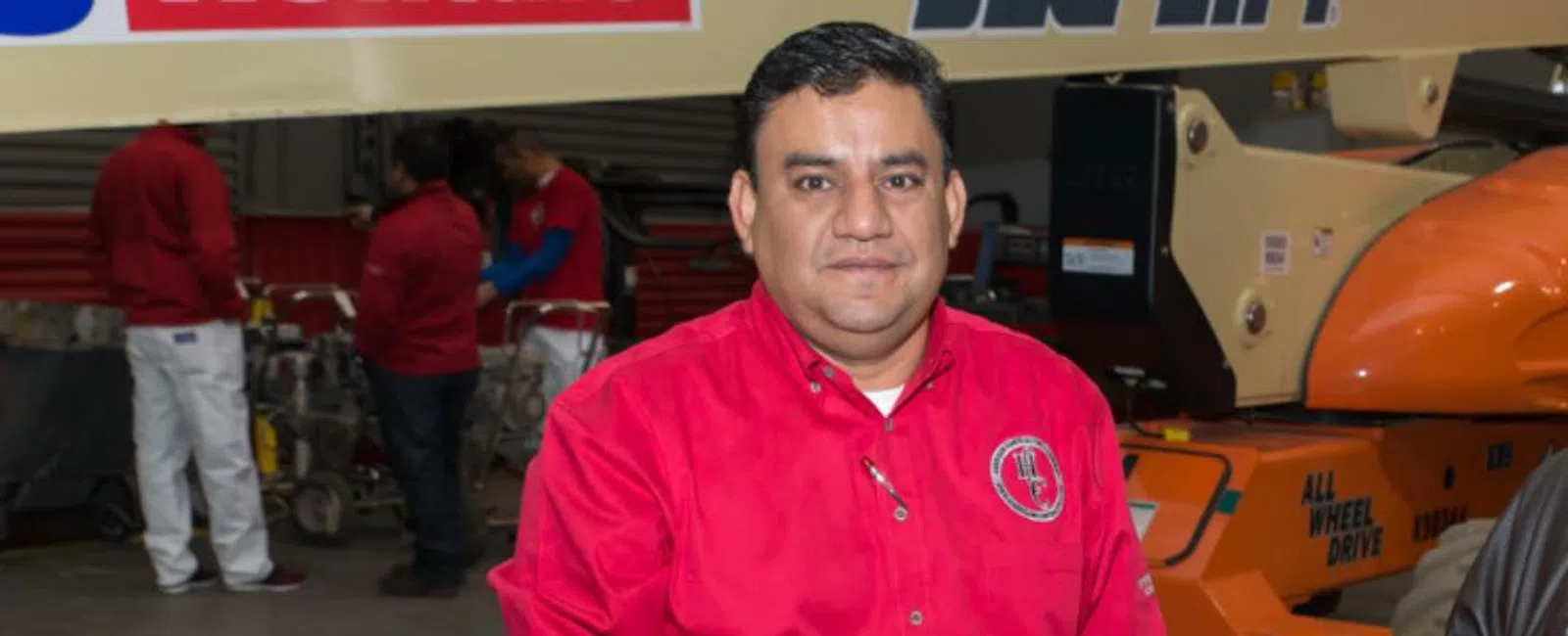 Man in red shirt and jeans holding a metal cup standing in an industrial warehouse with machinery.