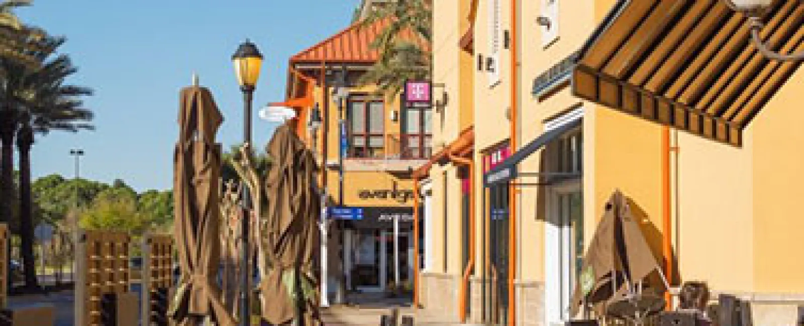 Sunny outdoor shopping plaza with yellow buildings, closed umbrellas, and empty patio tables under clear blue sky
