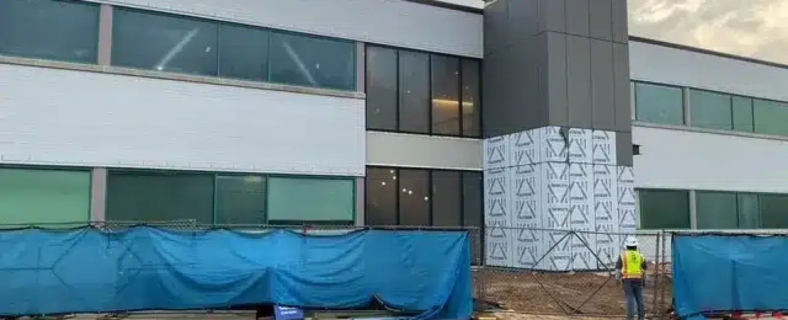 Modern commercial building under construction with worker and blue protective fencing at the site.