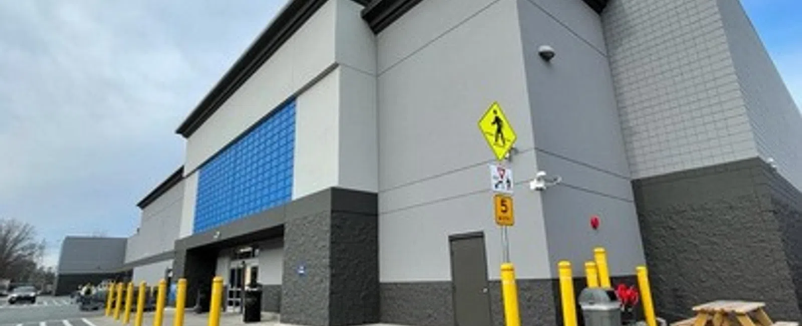 Large retail store exterior with gray walls, blue-tiled accent, yellow bollards, and pedestrian crossing sign under cloudy sky