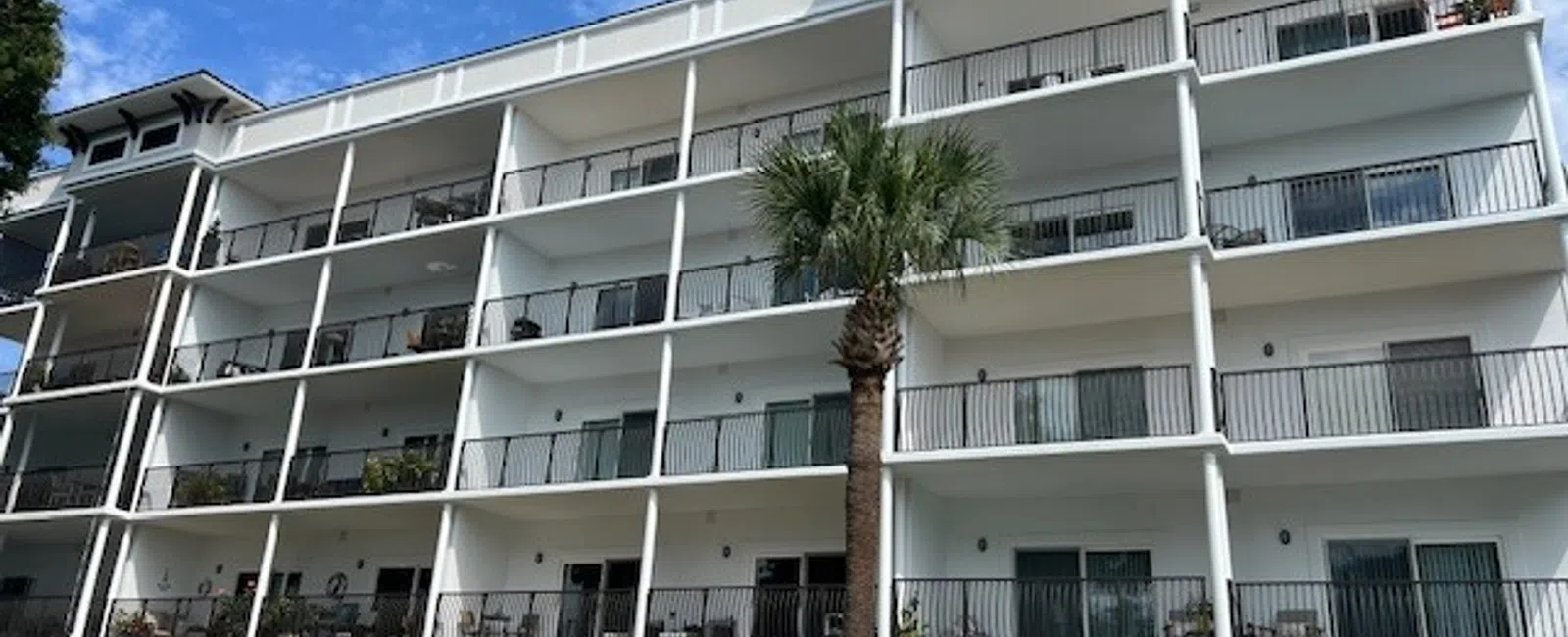 Modern white apartment building with balconies and a palm tree under a blue sky with clouds. Fall Maintenance Checklist for Facility Managers.