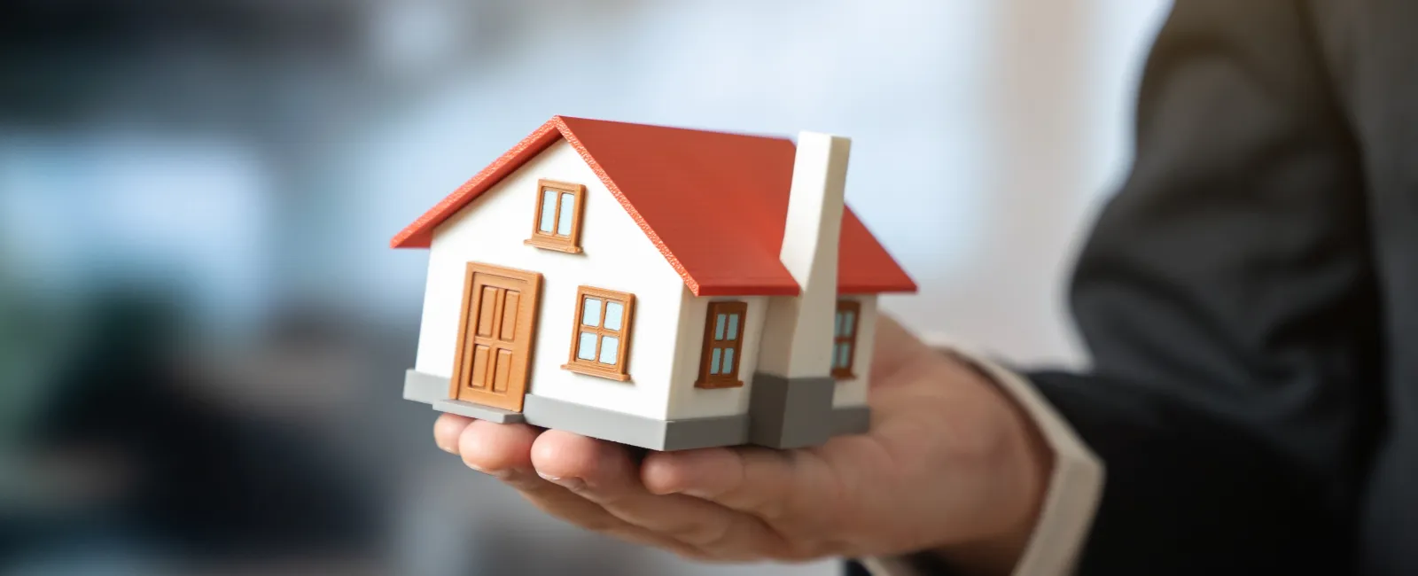 Hand holding a small model house with a red roof and brown door, representing real estate or home buying.