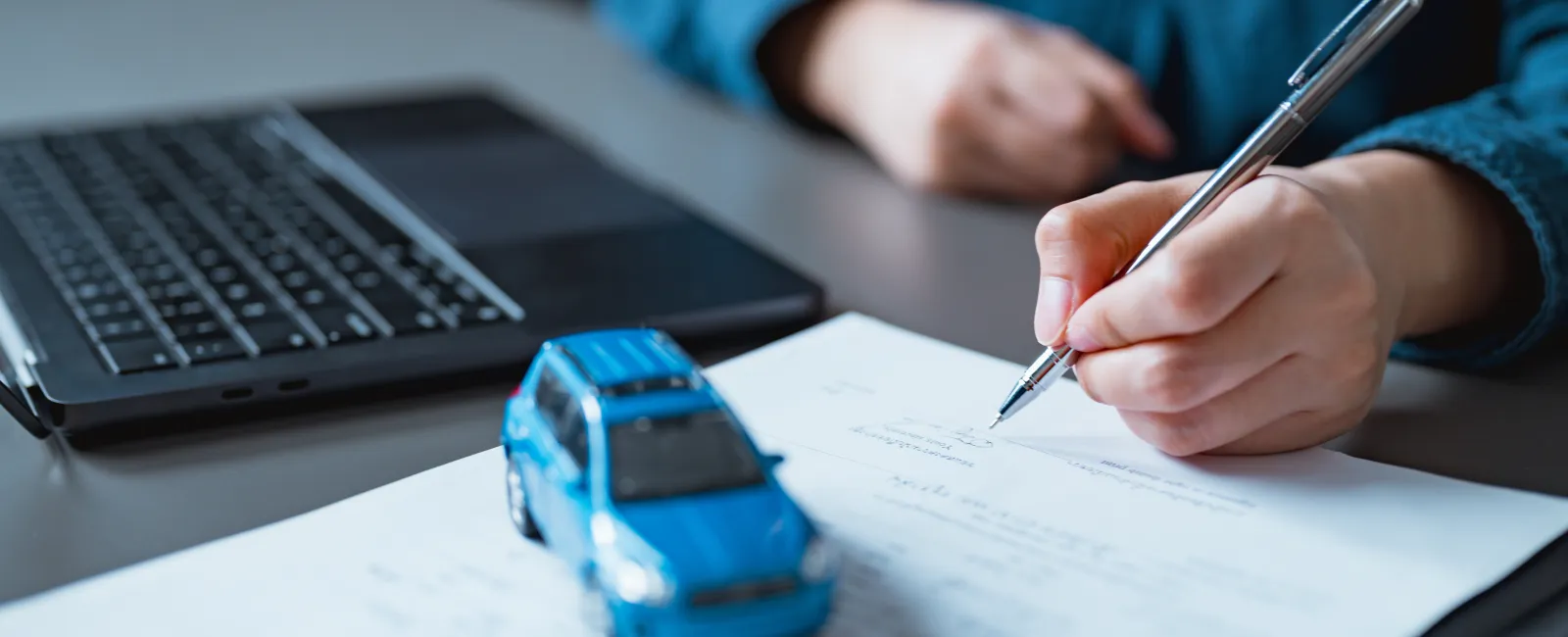 Person signing documents near a laptop and a blue toy car on a desk, symbolizing car insurance or purchase.