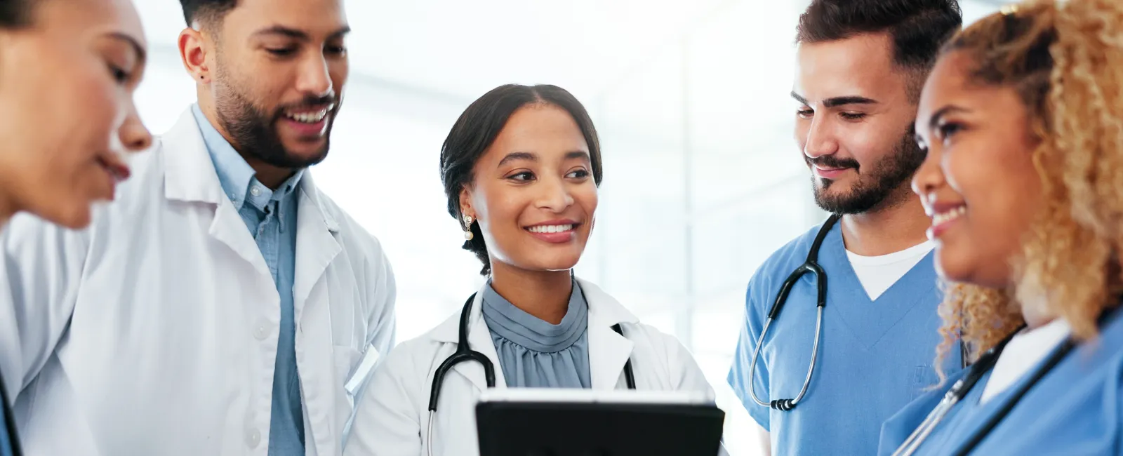 Diverse medical team in white coats and scrubs discussing patient information with a tablet in a bright hospital setting