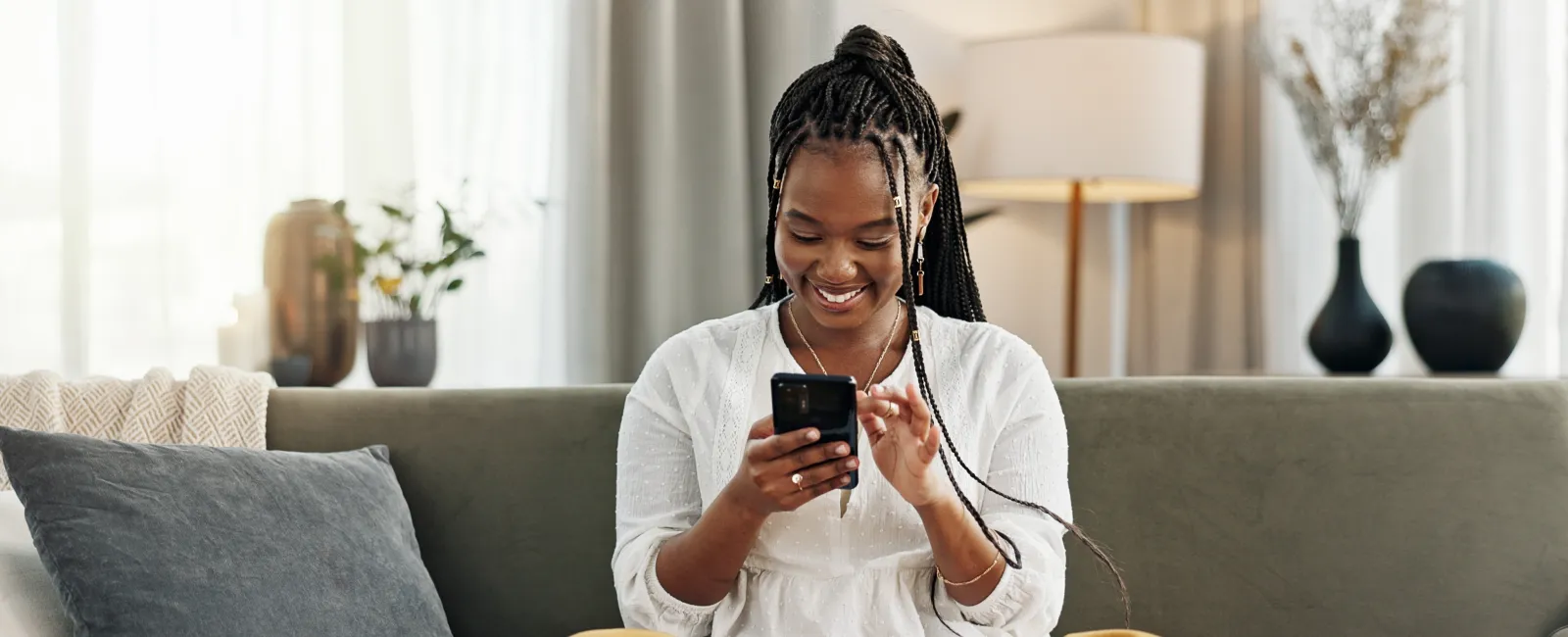 Happy young woman sitting cross-legged on couch using smartphone in cozy living room with laptop nearby
