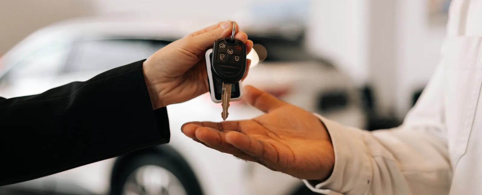 Person handing over car keys to another person with a white car in the background at a dealership.