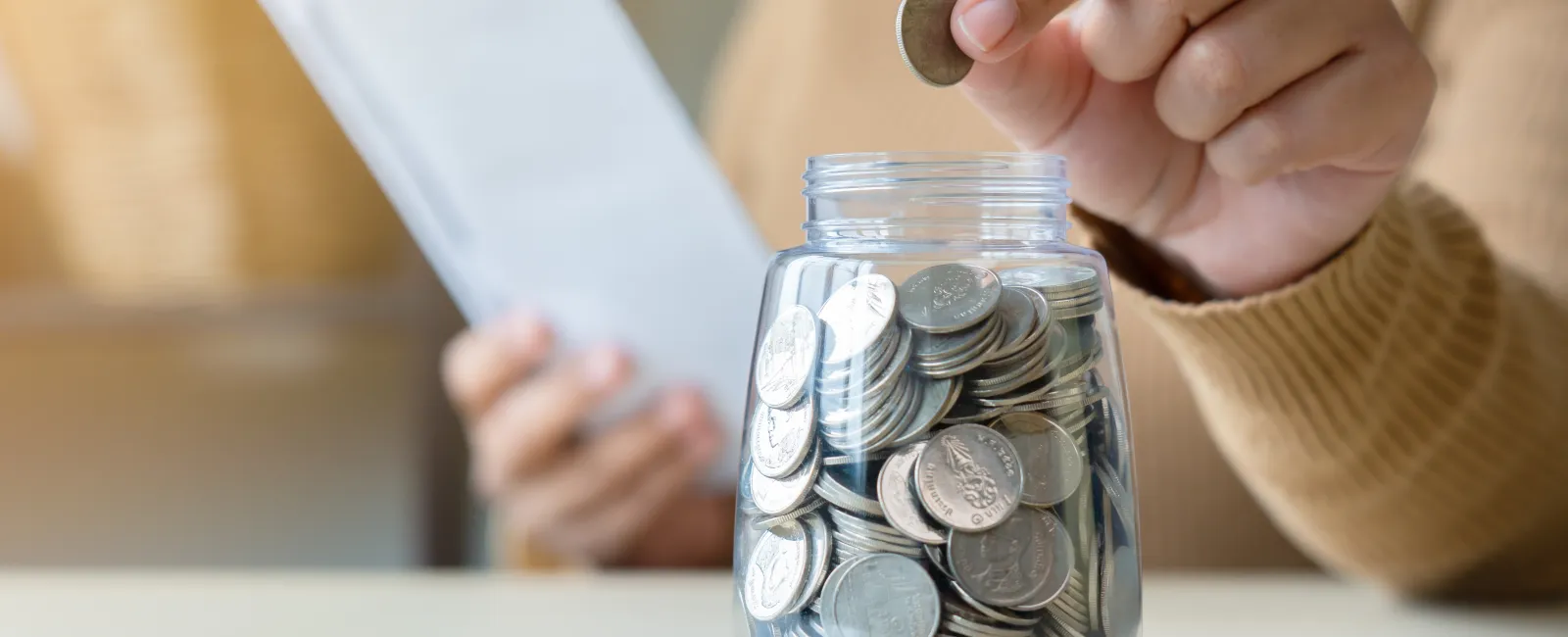 Person placing coin into jar filled with coins while holding a notebook, symbolizing saving money and budgeting.