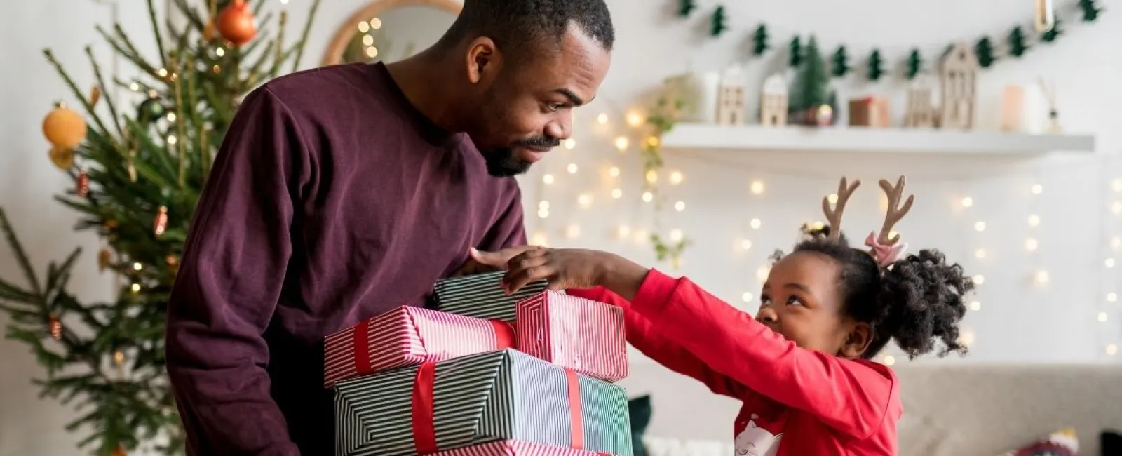 Father holding stack of Christmas gifts as happy daughter wearing reindeer antlers reaches for presents at home.