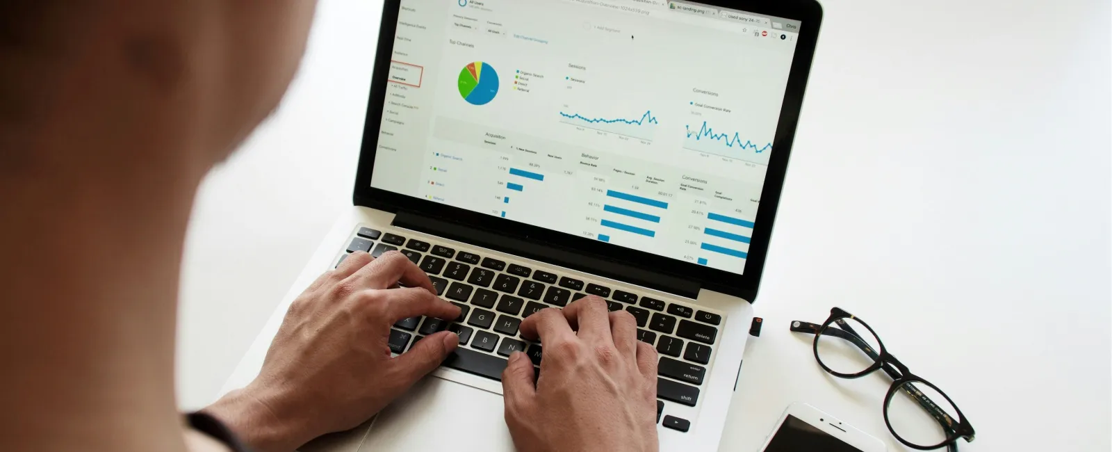 Person analyzing business data charts and graphs on a laptop with smartphone and glasses nearby on white desk