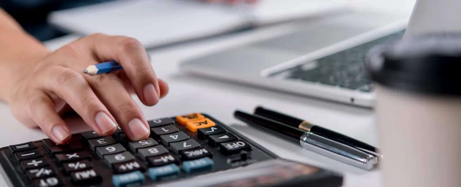 Close-up of hand using calculator with pen, laptop, and coffee cup on desk for work or study.