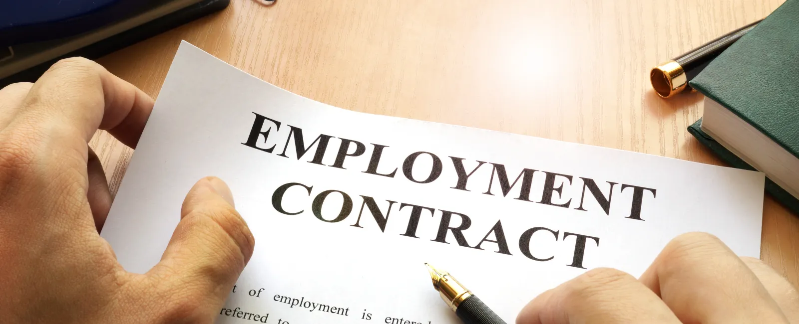 Person holding an employment contract document and a pen ready to sign on a wooden desk with office supplies.