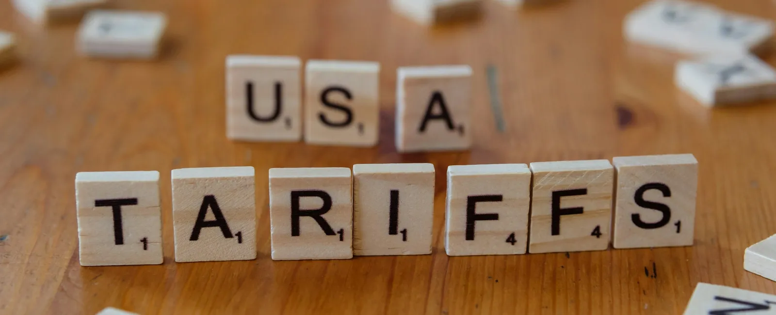 Wooden letter tiles spelling USA and TARIFFS on a wooden table with scattered tiles around.