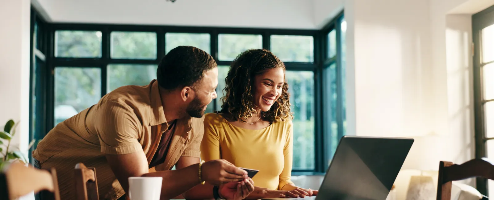 Smiling couple shopping online using laptop and credit card at a bright kitchen table.