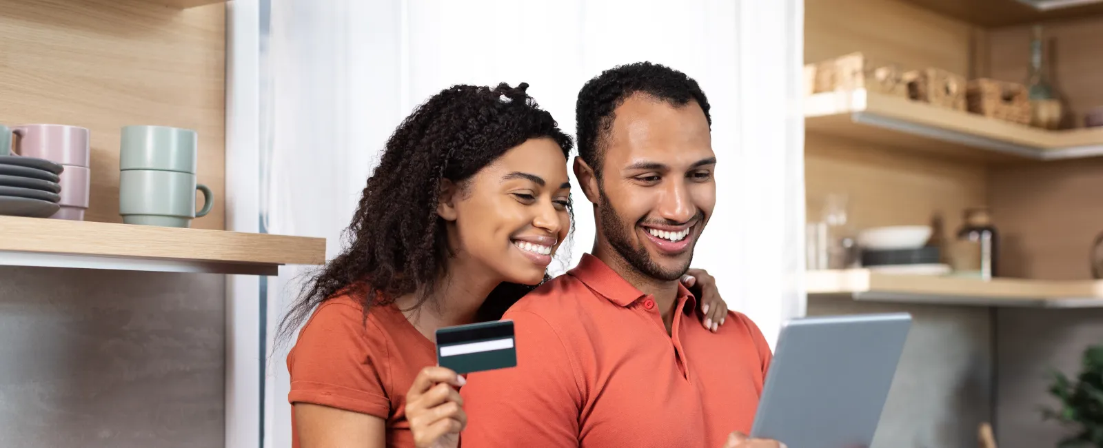 Happy couple in matching orange shirts shopping online with tablet and credit card in modern kitchen.