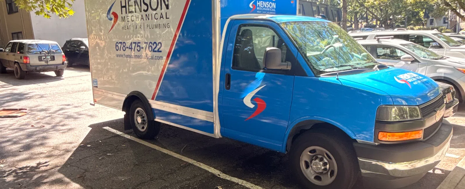 Blue and white Henson Mechanical service truck parked under leafy trees in a sunlit parking lot with contact details visible.