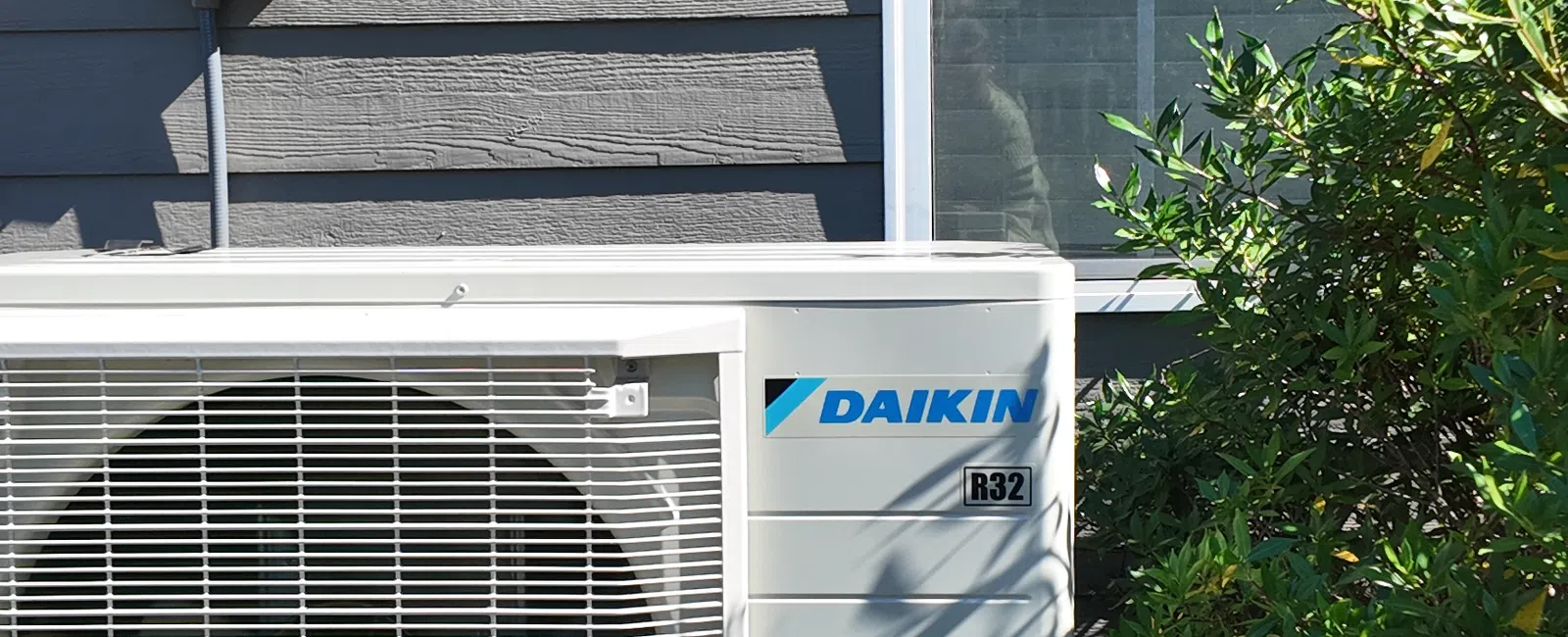 Daikin R32 air conditioning unit installed outside a gray house near a window and green shrubbery.